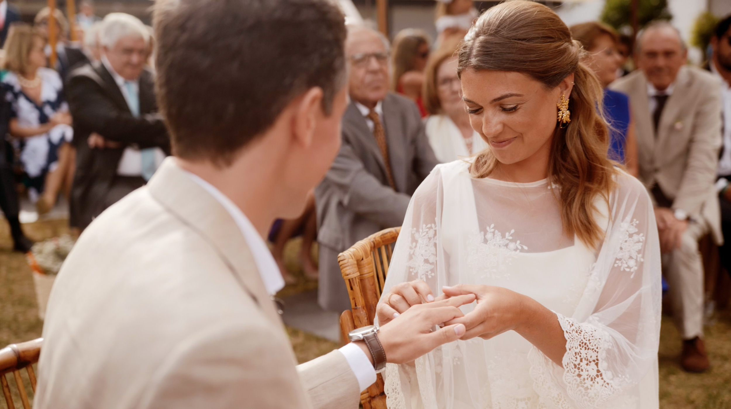 A wedding ceremony outdoors with a woman in a white lace dress exchanging rings with a man in a white suit, surrounded by seated guests.