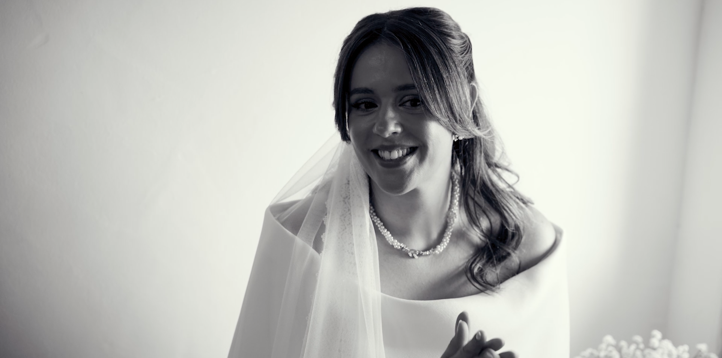 A woman smiling, wearing a wedding veil, pearl necklace, and white wedding dress, standing against a plain background.