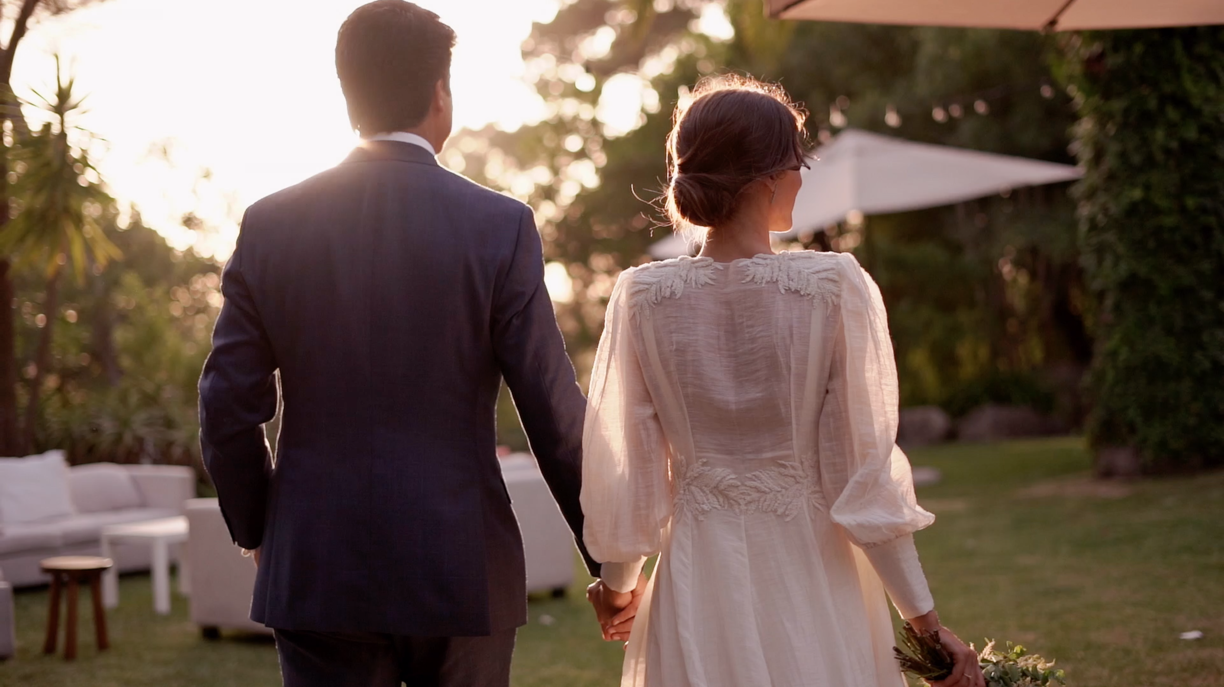 A bride and groom holding hands outdoors during an event at sunset, with trees and garden furniture in the background.