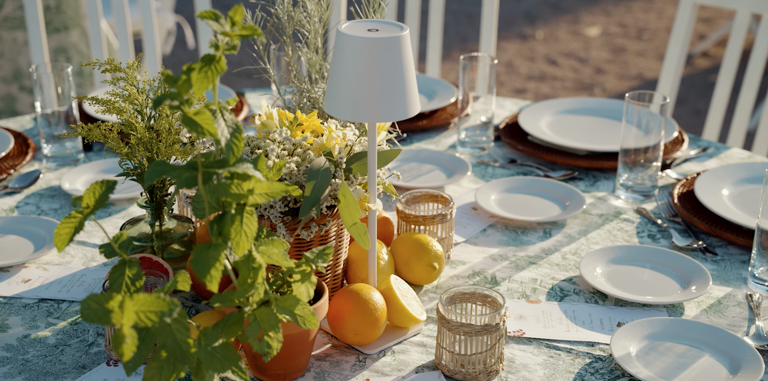 Table set for outdoor dining with white plates, clear glasses, silverware, a floral centerpiece with white and yellow flowers, lemons, and greenery, on a patterned tablecloth.