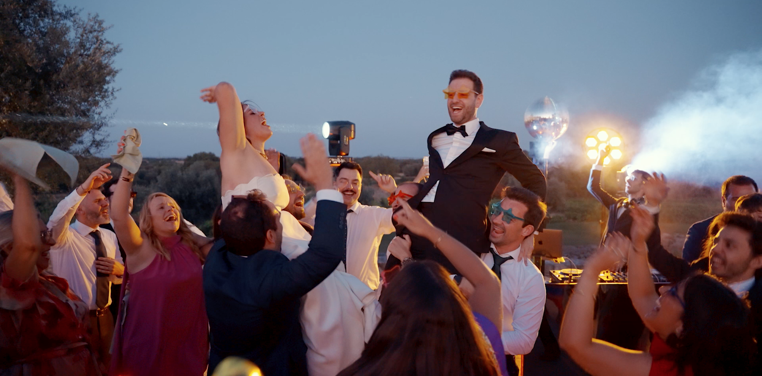 People celebrating outdoors at sunset, lifting a man dressed in a tuxedo and sunglasses in the air during a party or wedding celebration.