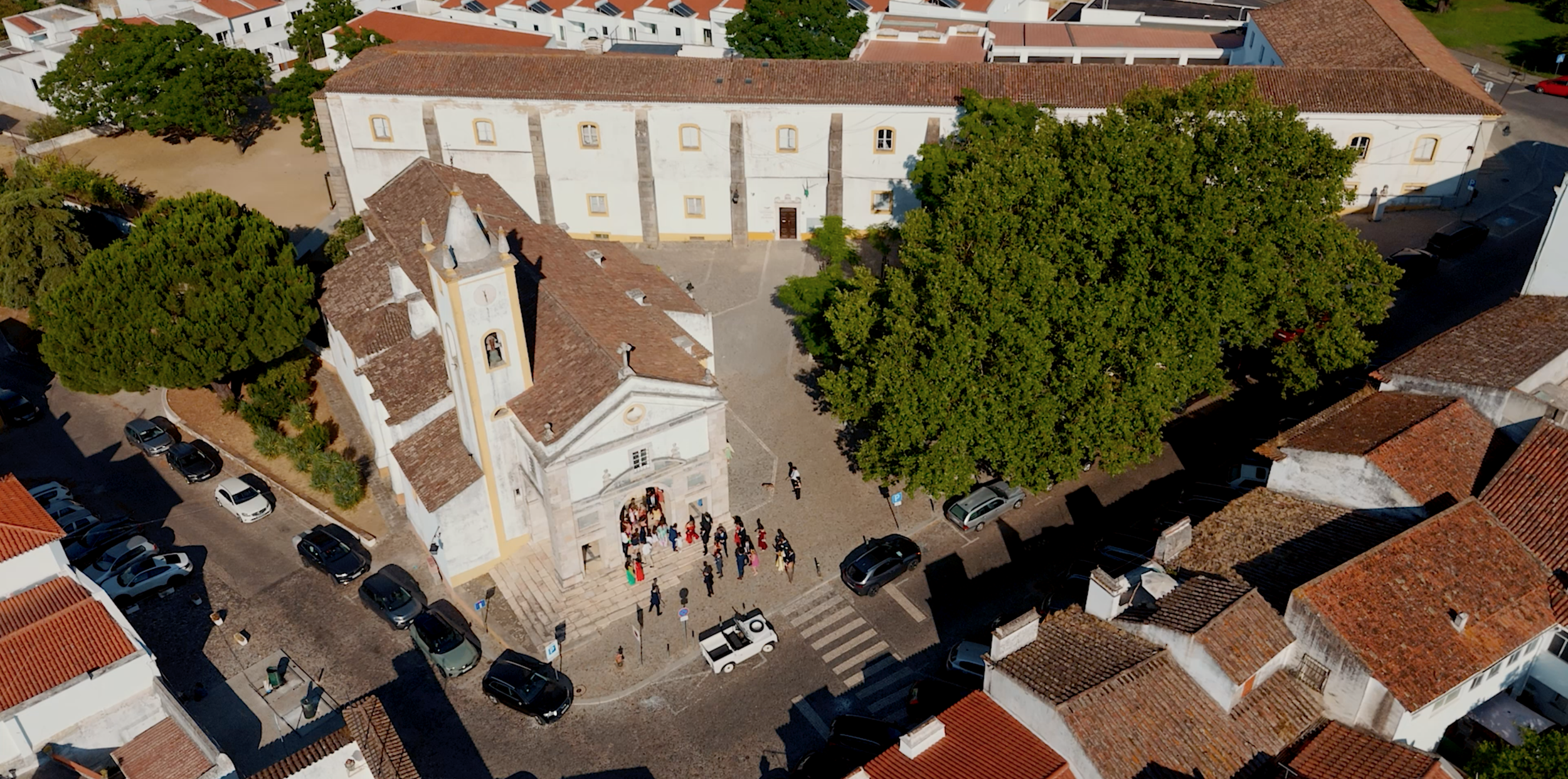 Aerial view of a historic church with a small crowd gathered at the entrance, surrounded by trees, parked cars, and nearby buildings with tiled roofs.