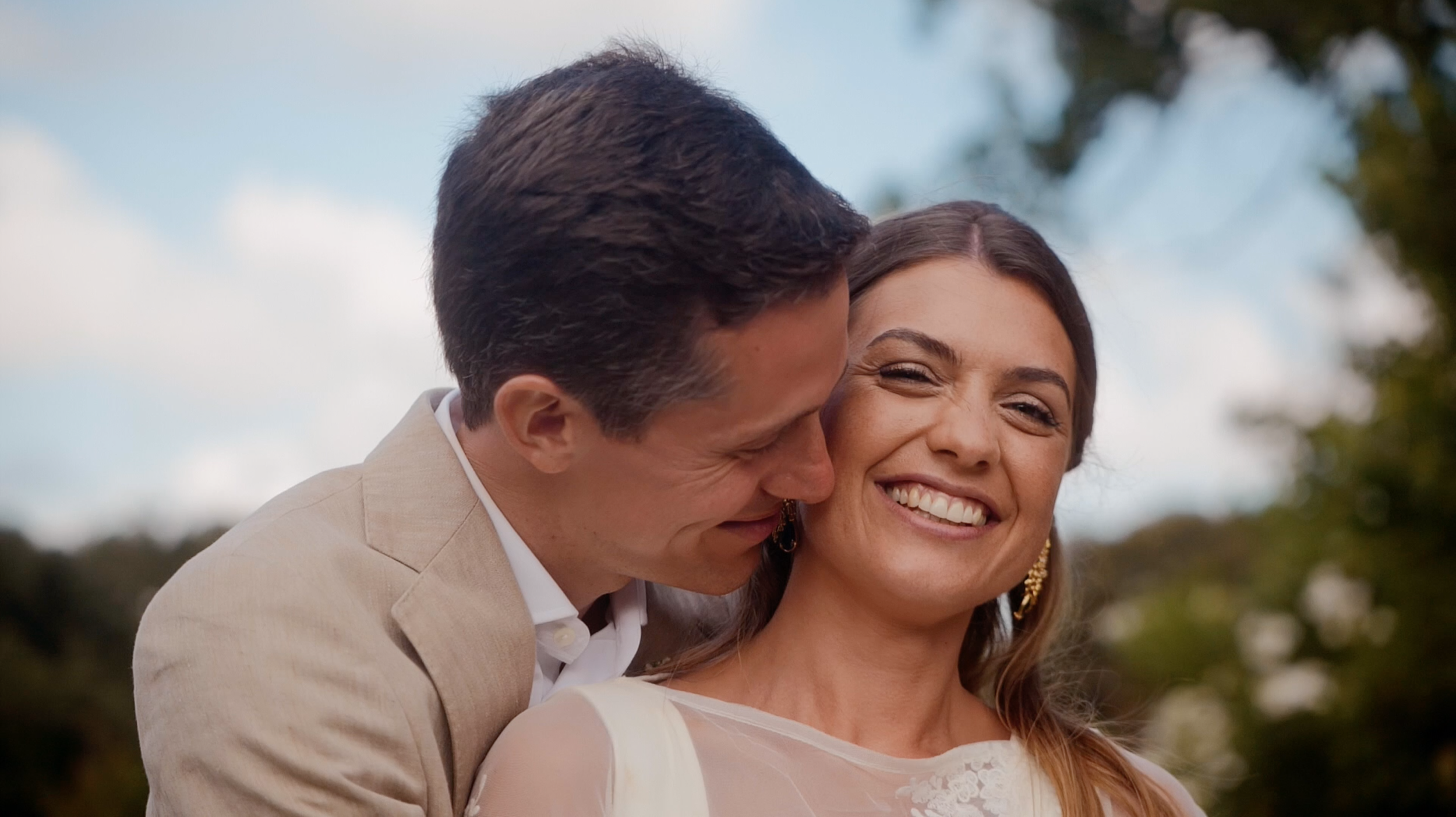 A man and woman smiling and sharing a close moment outdoors, with a blue sky and trees in the background.