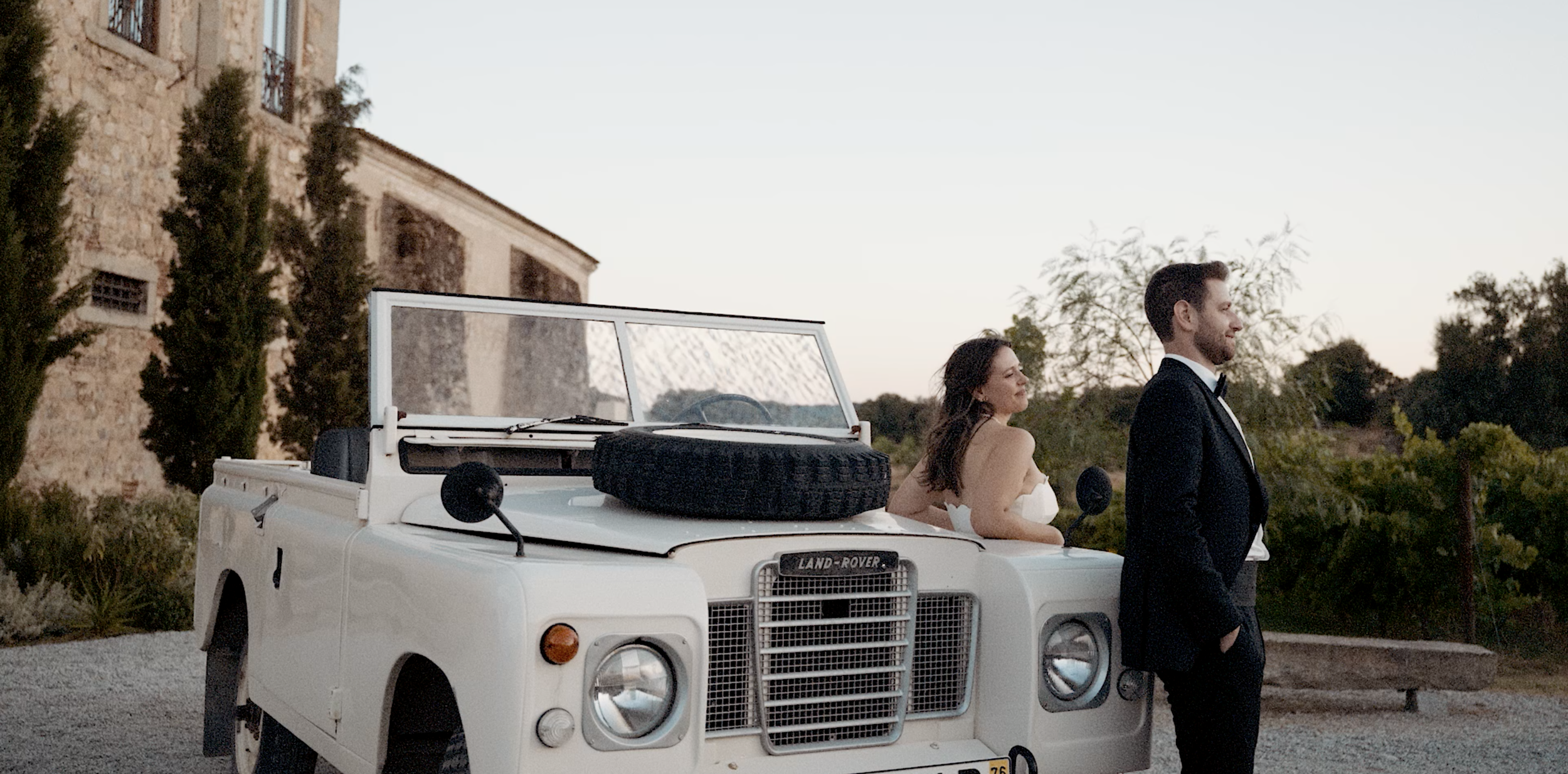 A woman in a white dress and a man in a tuxedo standing near a white vintage Land Rover with a spare tire on the hood, in front of an old stone building with trees in the background.