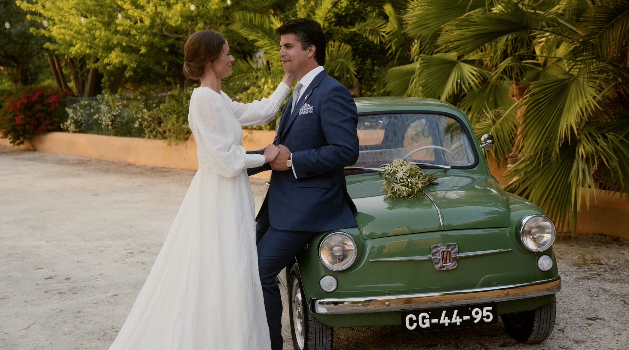 A bride and groom holding hands, smiling at each other, standing outside near a vintage green Fiat car decorated with flowers, with lush green trees and bushes in the background.