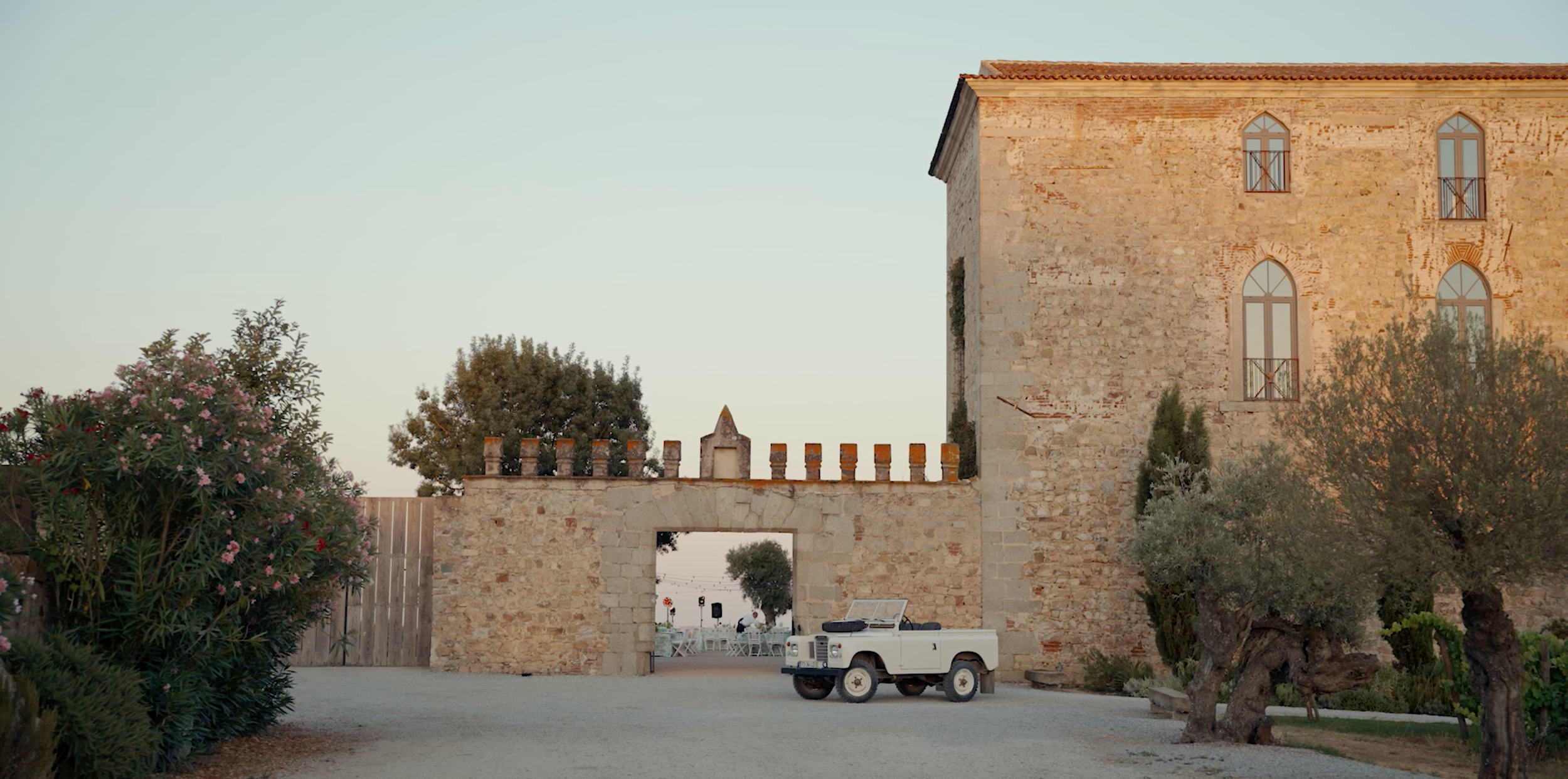A stone building with tall arched windows and a red tile roof. There are trees and flowers in the foreground, a white utility vehicle parked, and a wall with decorative crenellations.