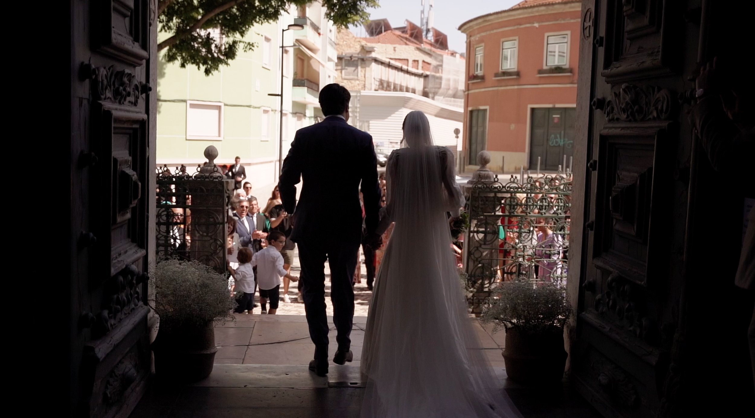 A bride and groom holding hands at the entrance of a church, facing a crowd of wedding guests, with city buildings in the background.