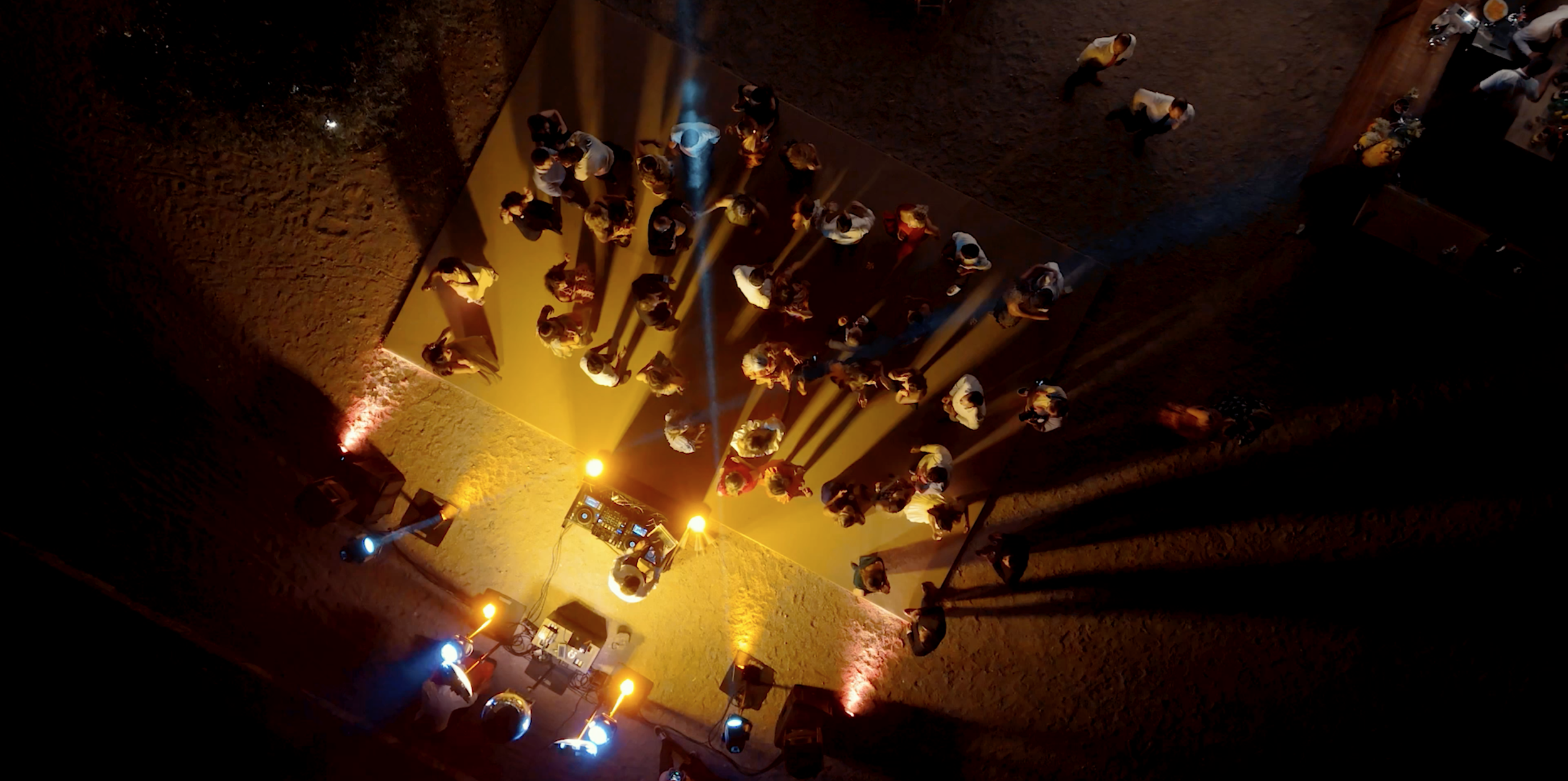 A bird's eye view of a dance floor with people dancing, illuminated by yellow and blue stage lights in a rustic stone-walled venue.