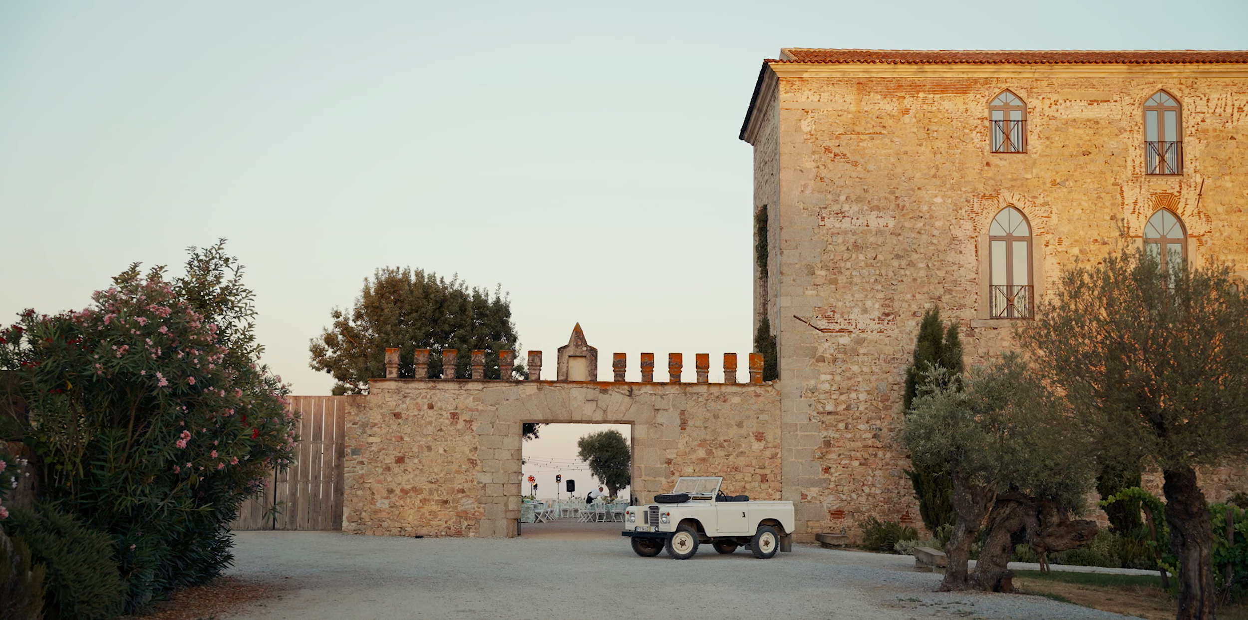 A rustic stone building with arched windows and a red tile roof, surrounded by trees and plants, with a vintage white vehicle parked in front.