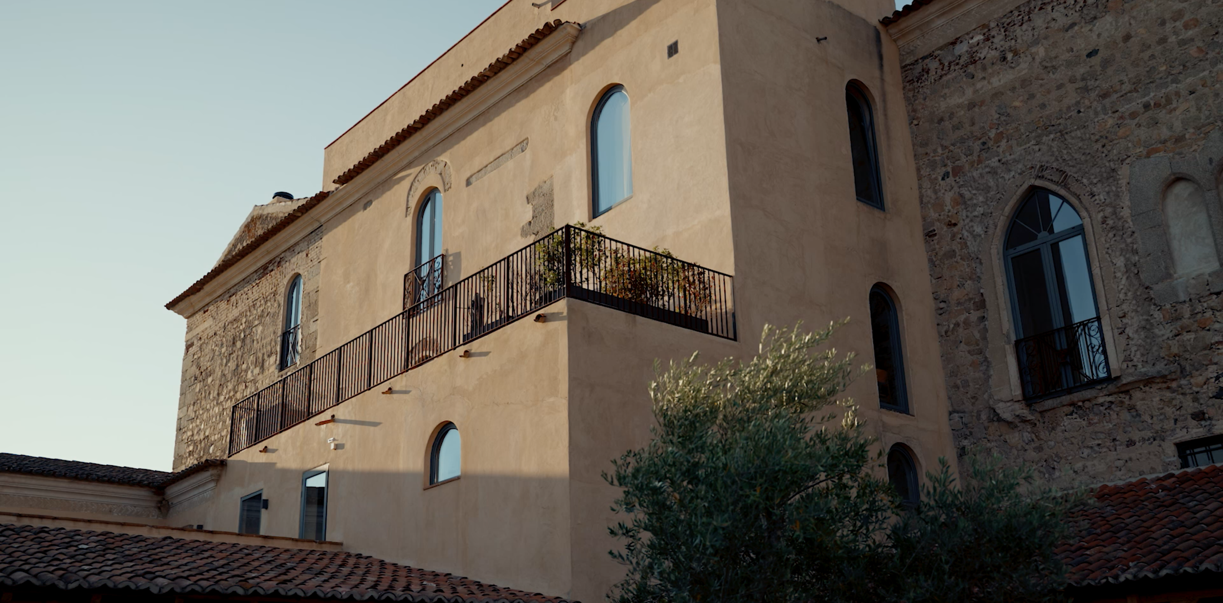 A beige and stone building with arched windows, a balcony with a black railing, and a tiled roof, under a clear sky.