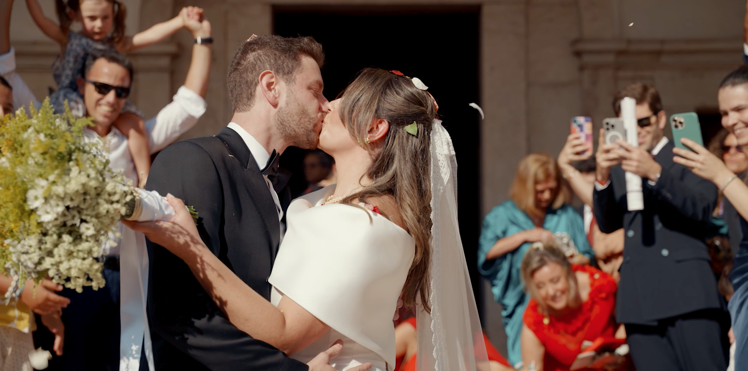 A bride and groom kissing during their wedding ceremony surrounded by family and friends, some of whom are taking photos. The bride is holding a bouquet of flowers, and the groom is dressed in a tuxedo.