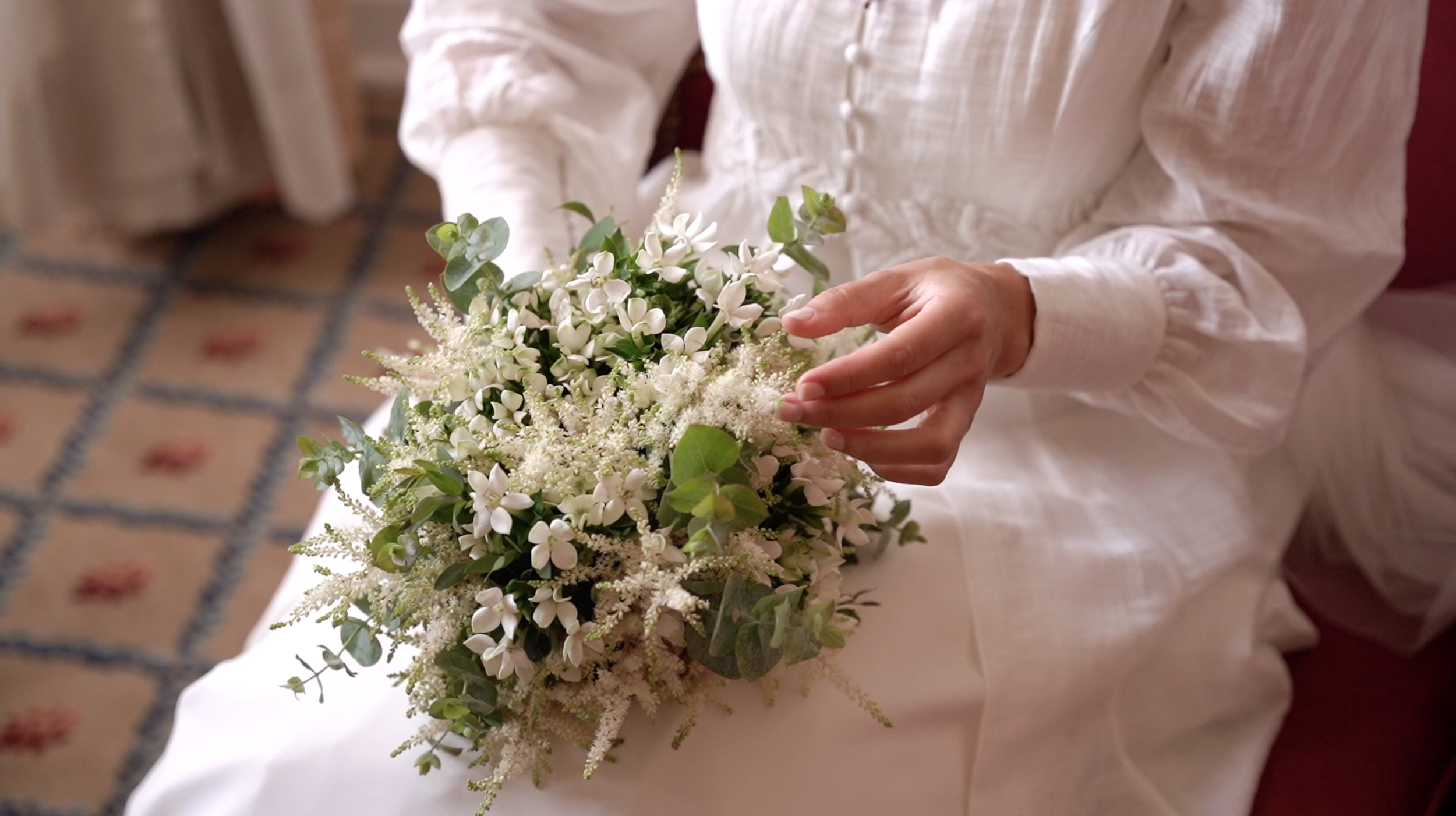 Person holding a bouquet of white flowers with green leaves, wearing a long-sleeved white garment, in an indoor setting with a patterned tile floor.