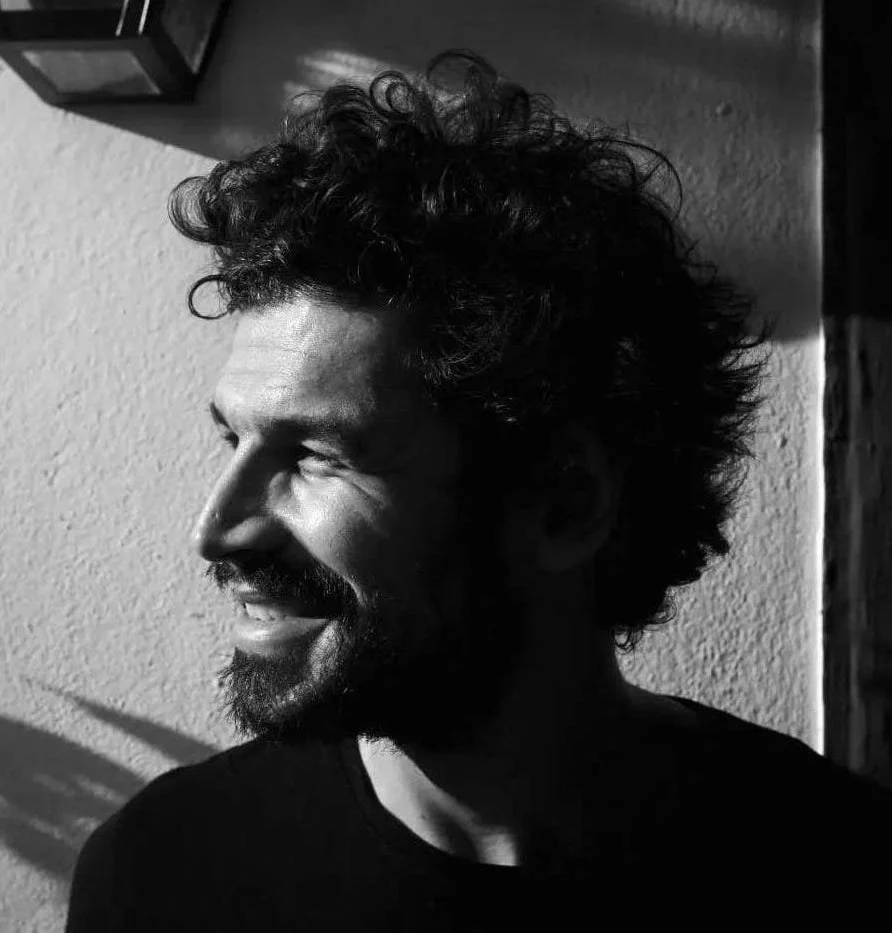 Black and white photo of a smiling man with curly hair and a beard, looking to the left, with light casting shadows on the textured wall behind him.
