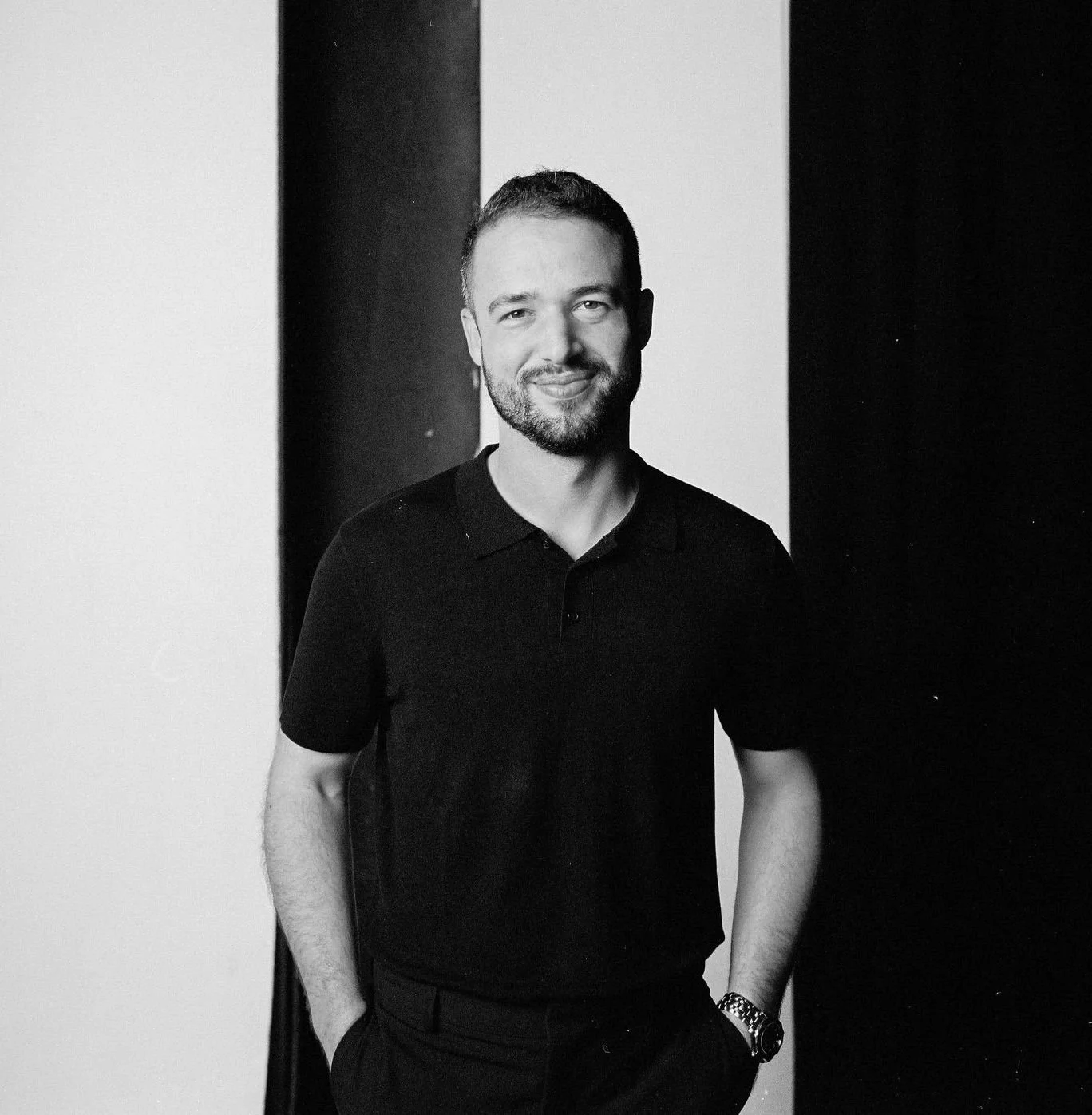 A black and white photo of a man with a short beard and short hair, smiling and standing with one hand in his pocket, wearing a dark polo shirt, against a background with vertical black and white stripes.
