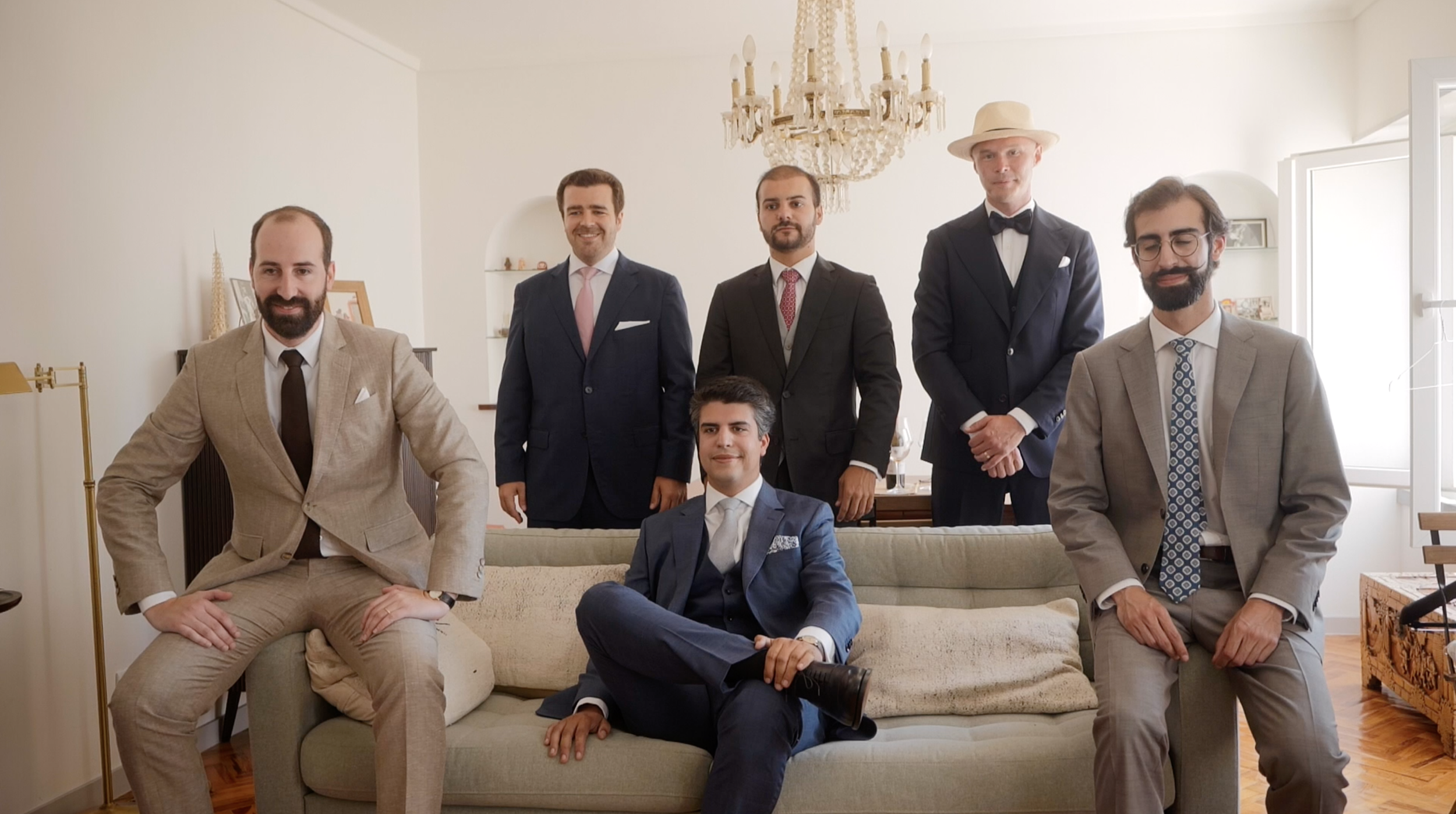 A group of seven men in suits, some standing and some seated, in a bright living room with a chandelier overhead and artwork in the background.