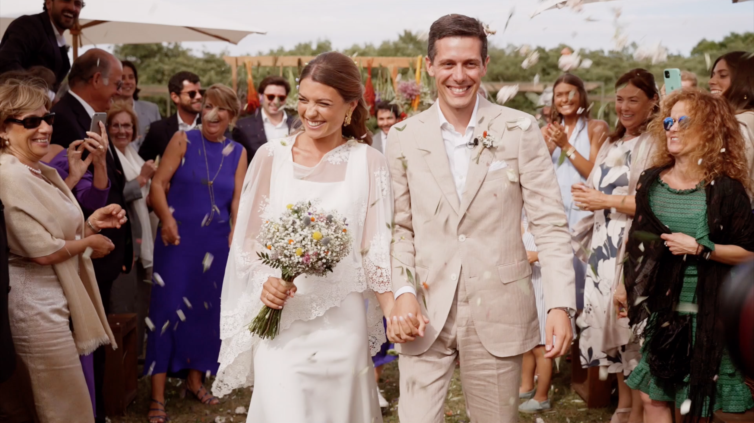 A newlywed couple walking hand in hand at their outdoor wedding reception, surrounded by friends and family celebrating with smiles and throwing flower petals.