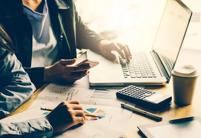 Two people working at a desk with a laptop, smartphone, calculator, documents, a pen, a tablet, and a coffee cup.