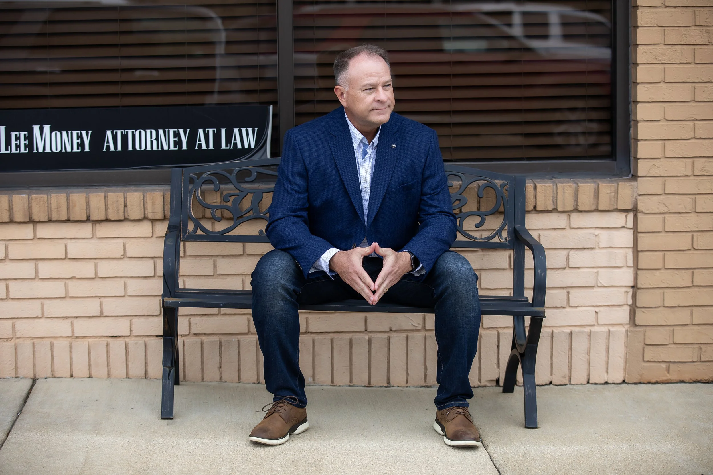 A man sitting on a black bench outside a building with a sign that reads 'Lee Money Attorney at Law'. He is wearing a blue blazer, light-colored shirt, dark jeans, and brown shoes, with hands clasped together.