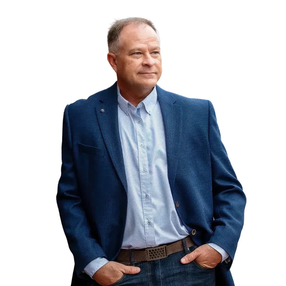 A middle-aged man with short hair, wearing a blue blazer and a light blue button-up shirt, standing with his hands in his pockets against a black background.