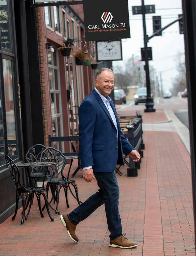 A man in a blue blazer and jeans walking on a brick sidewalk in a small downtown area, smiling, with outdoor tables and chairs in front of a building with business signs hanging above.
