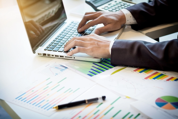 Person in business attire working on a laptop at a desk with financial charts, graphs, and a pen.