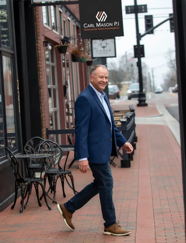 A man in a blue blazer and jeans walking on a brick sidewalk in a small downtown area, smiling, with outdoor tables and chairs in front of a building with business signs hanging above.
