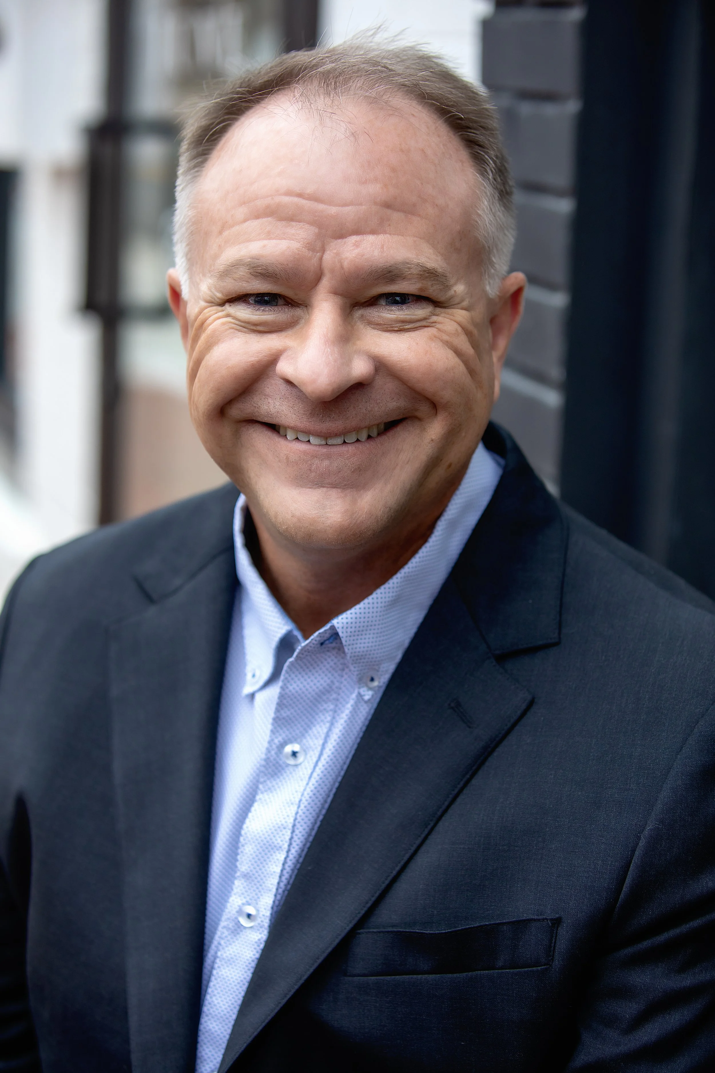 A smiling man wearing a black suit jacket, light blue shirt, and no tie, standing outdoors with a blurred background of a building or house.