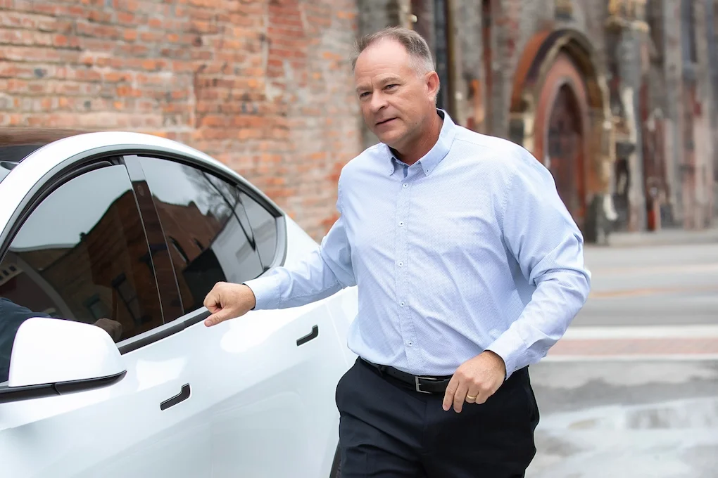 A middle-aged man in a light blue dress shirt and black pants standing next to a white car, with a brick building in the background.
