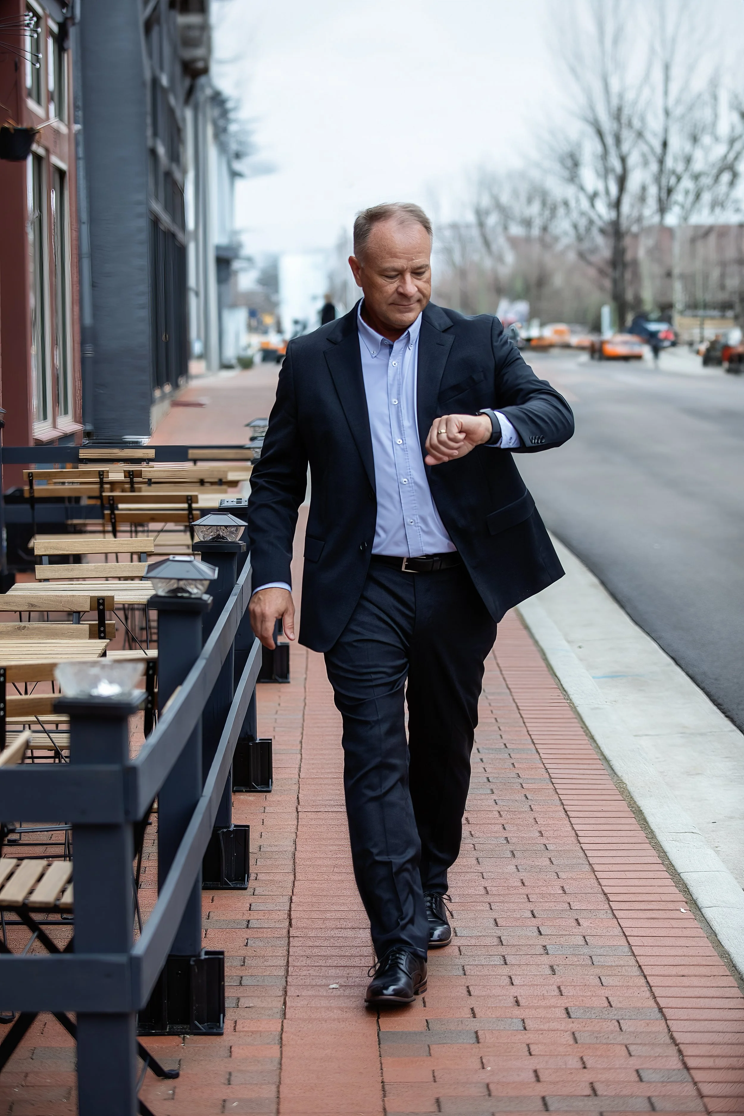 A man in a dark navy suit and light blue shirt walking on a brick sidewalk while looking at his watch. The sidewalk is lined with outdoor seating and small lamps, with buildings and leafless trees in the background.