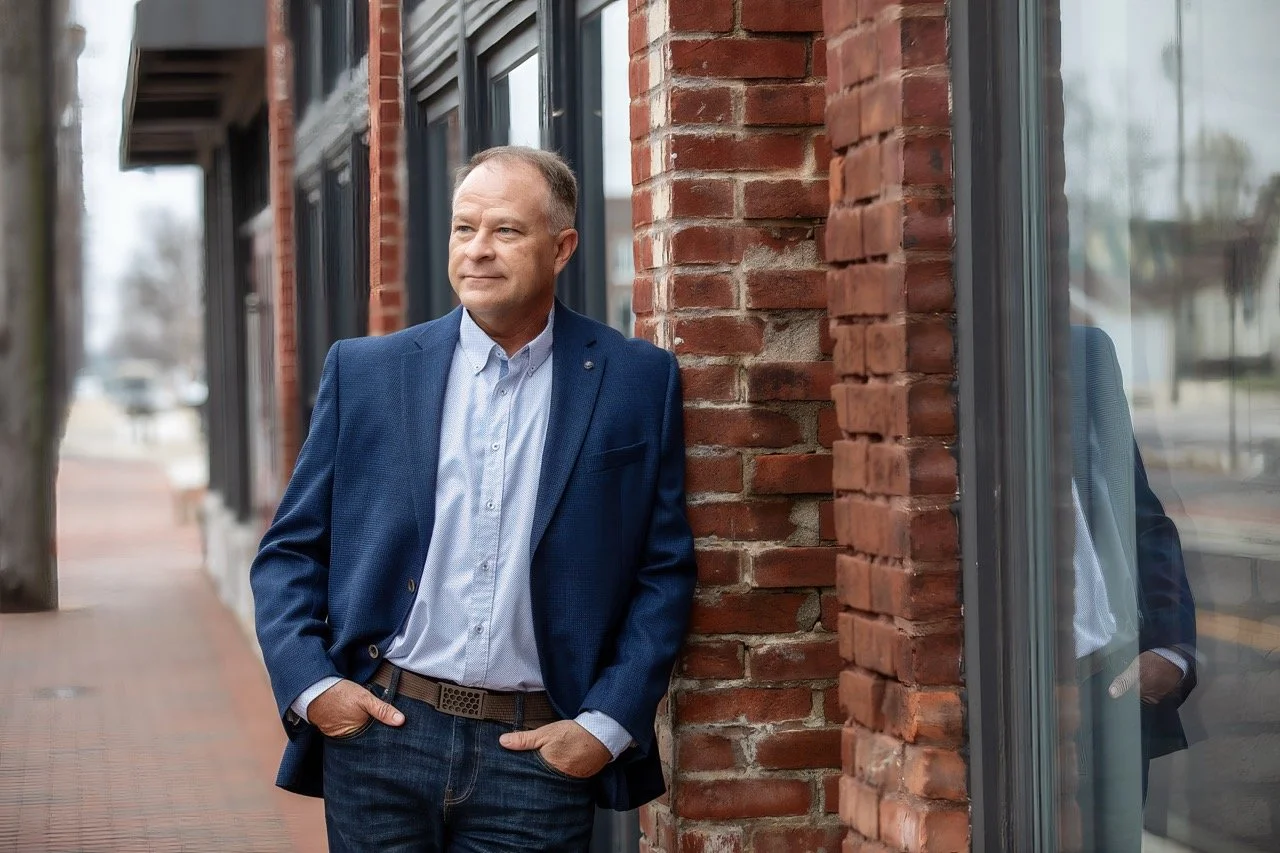 A man in a blue blazer and light blue shirt stands outside next to a brick building, with his hands in his pockets and looking off into the distance.