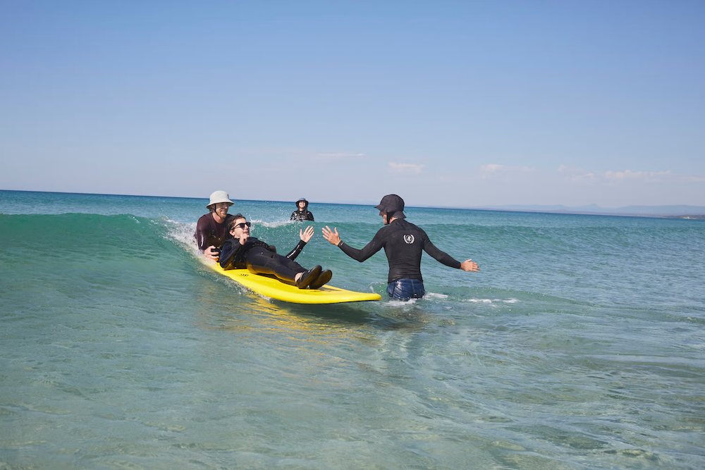 People at the beach helping a person on a yellow surfboard in the water.
