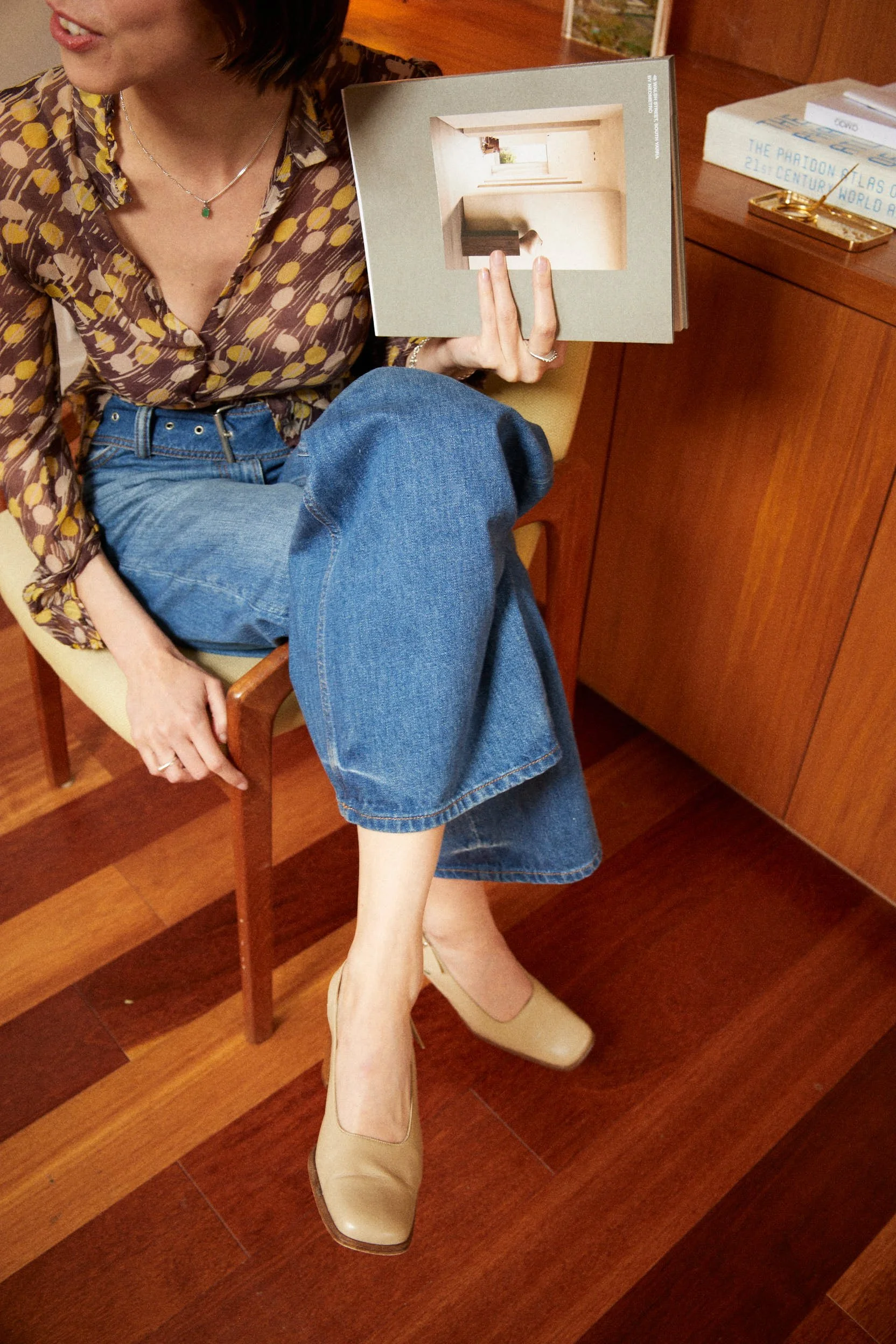 A woman sitting on a wooden chair, wearing a patterned blouse and blue jeans, holding a photo book while showing her legs to reveal beige high heels and part of her face.