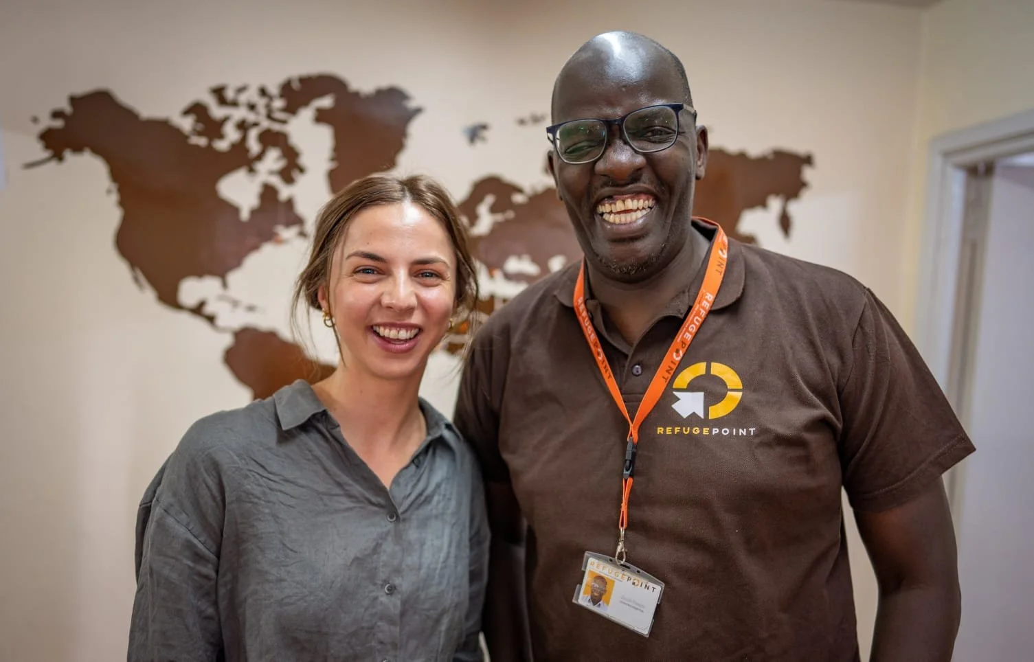 Two smiling employees, a woman and a man, standing in front of a world map wall decor at Refuge Point, wearing company shirts and ID badges.