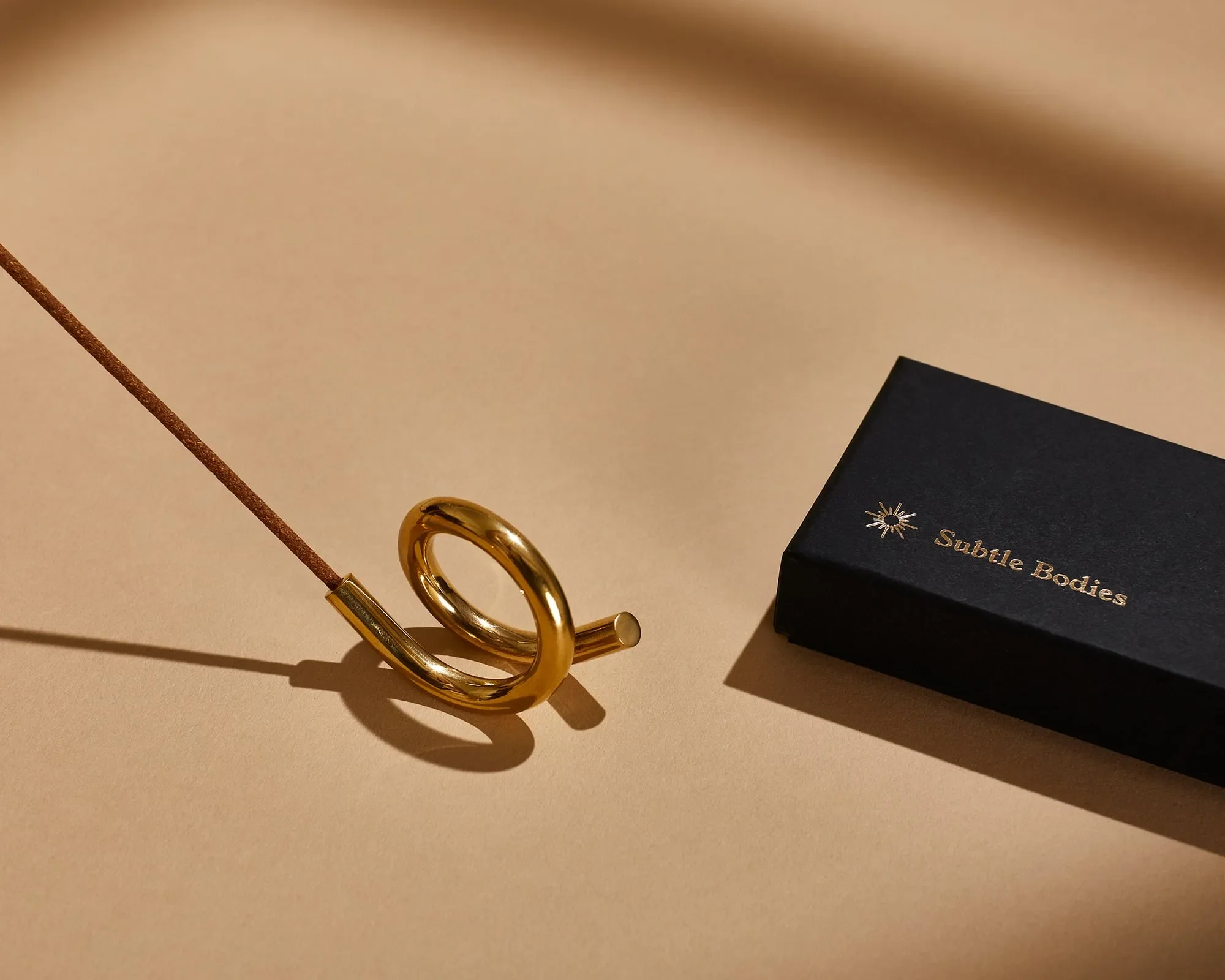 A gold ring, a black box with a starburst design and the words "Subtle Bodies," a gold curved object, and a copper wire on a beige surface.