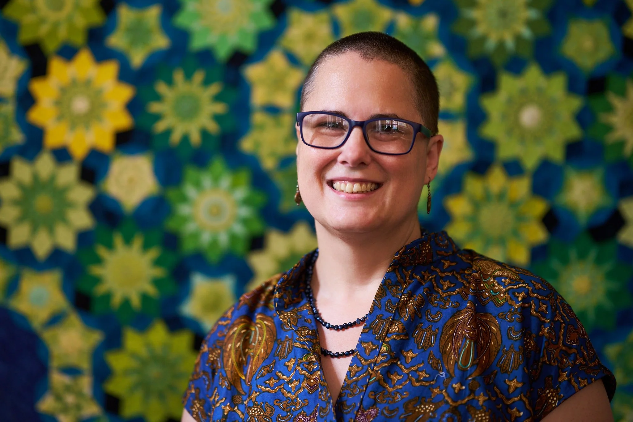 Portrait of Lynn smiling at the camera with a yellow and green and teal quilt in the background. Lynn is a white woman with a shaved head, blue glasses and a blue batik top.