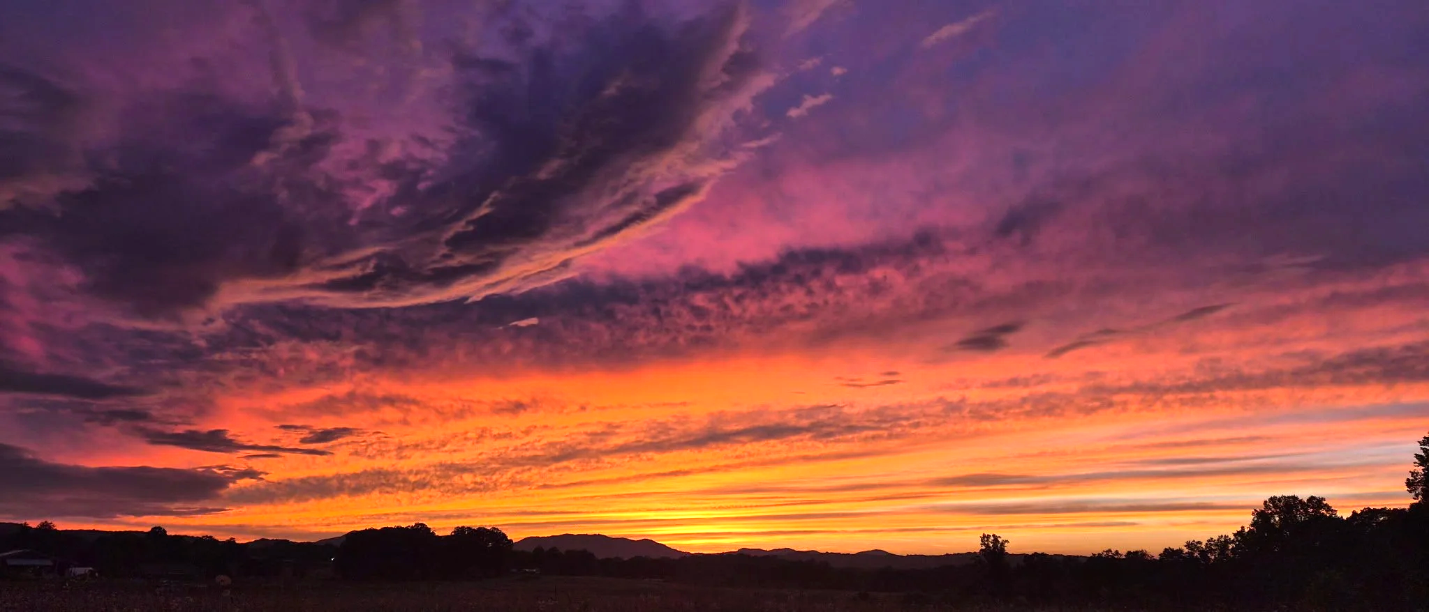 Colorful sunset sky with vibrant pink, purple, and orange clouds over a rural landscape with trees and distant mountains.