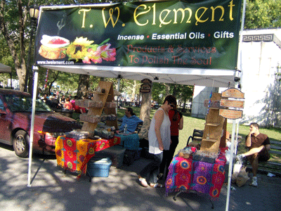 Outdoor market stall for T. W. Element, selling essential oils, gifts, and polishes, decorated with colorful tablecloths, under a green canopy with signage.