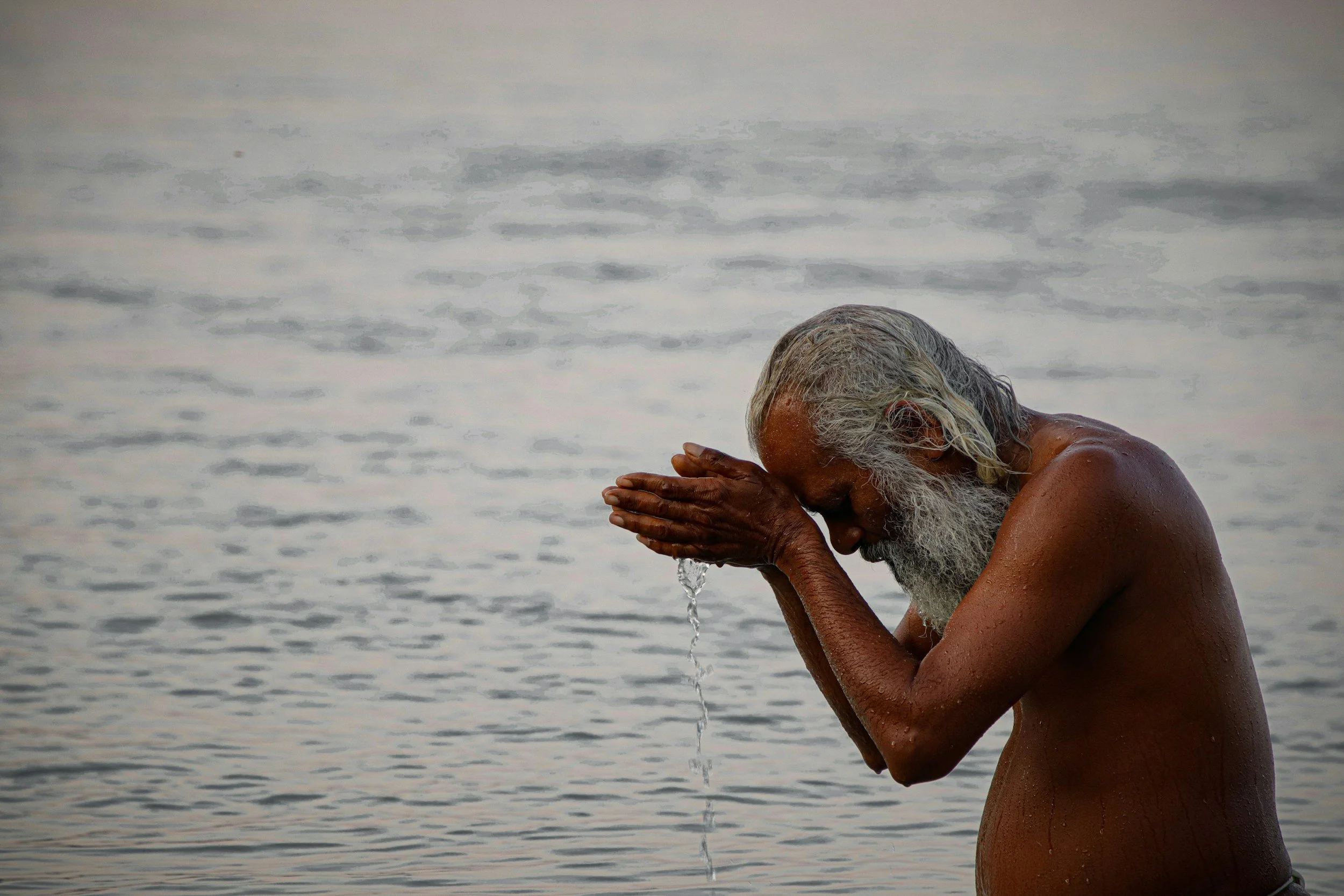An elderly man with long white hair and a beard washing his face in a body of water, possibly at the beach, with water droplets on his hands and body.