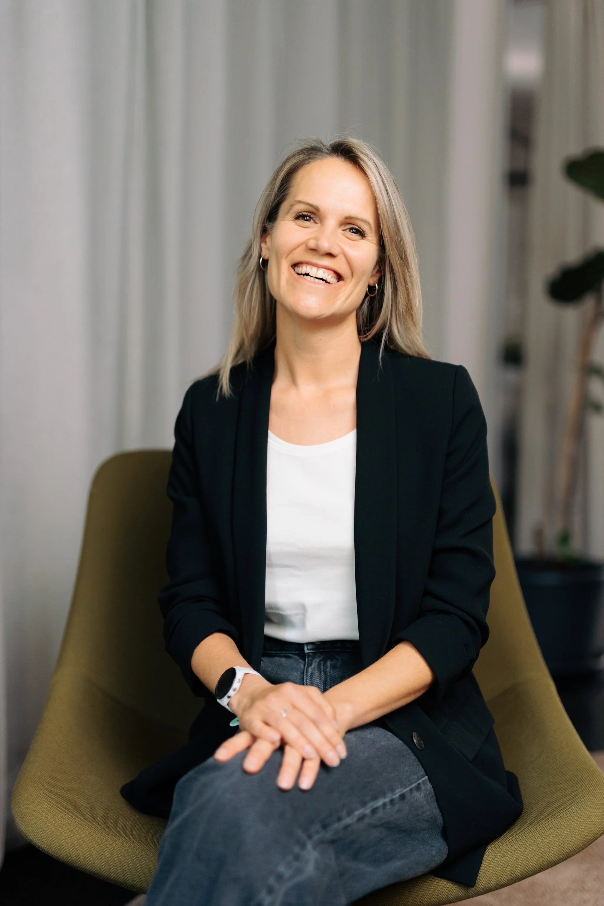 Eine Frau mit langem, blondem Haar lächelt, sitzt in einem grünen Stuhl, trägt einen schwarzen Blazer, ein weißes Shirt und eine dunkle Jeans, in einem professionellen Umfeld.