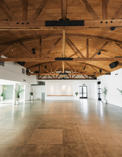 Spacious, empty event hall with wooden ceiling, white walls, and large floor, decorated with potted plants along the sides.