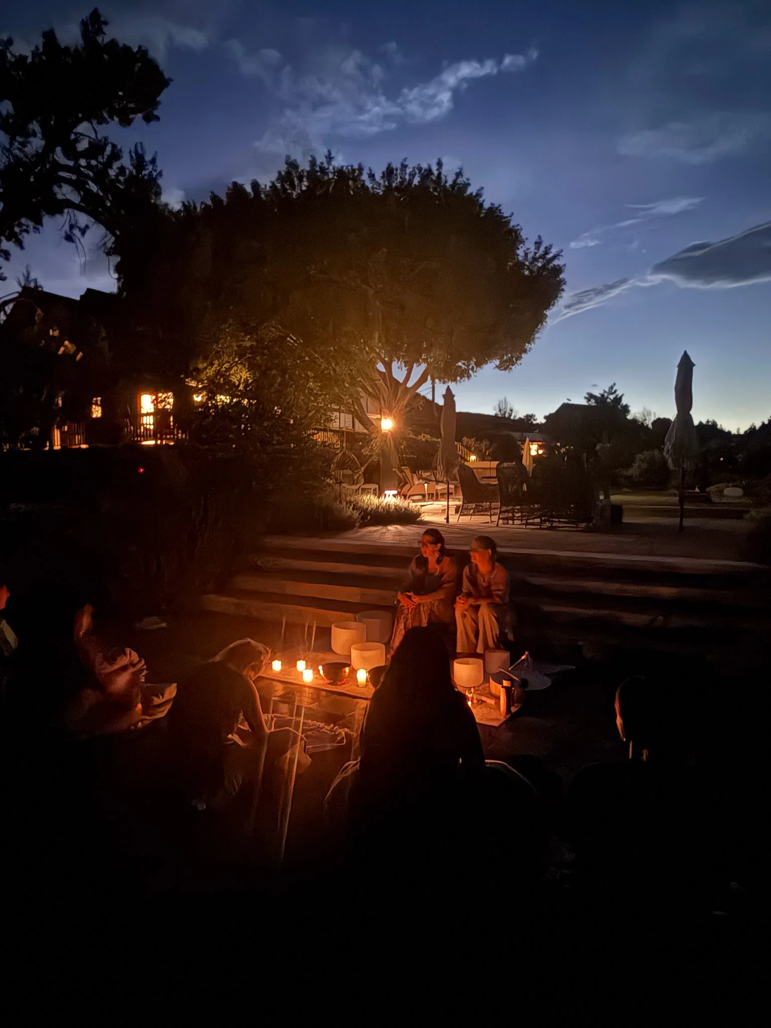 People gathered outdoors at night around a small fire, with candles and lanterns, under trees with a house in the background.