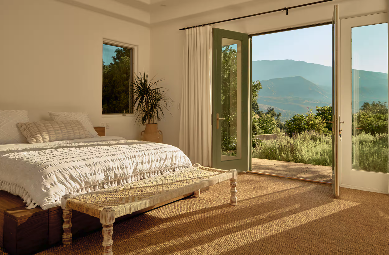 A bedroom with a large bed, a large potted plant, and open glass doors leading to a balcony with a view of green trees and mountains in the background.