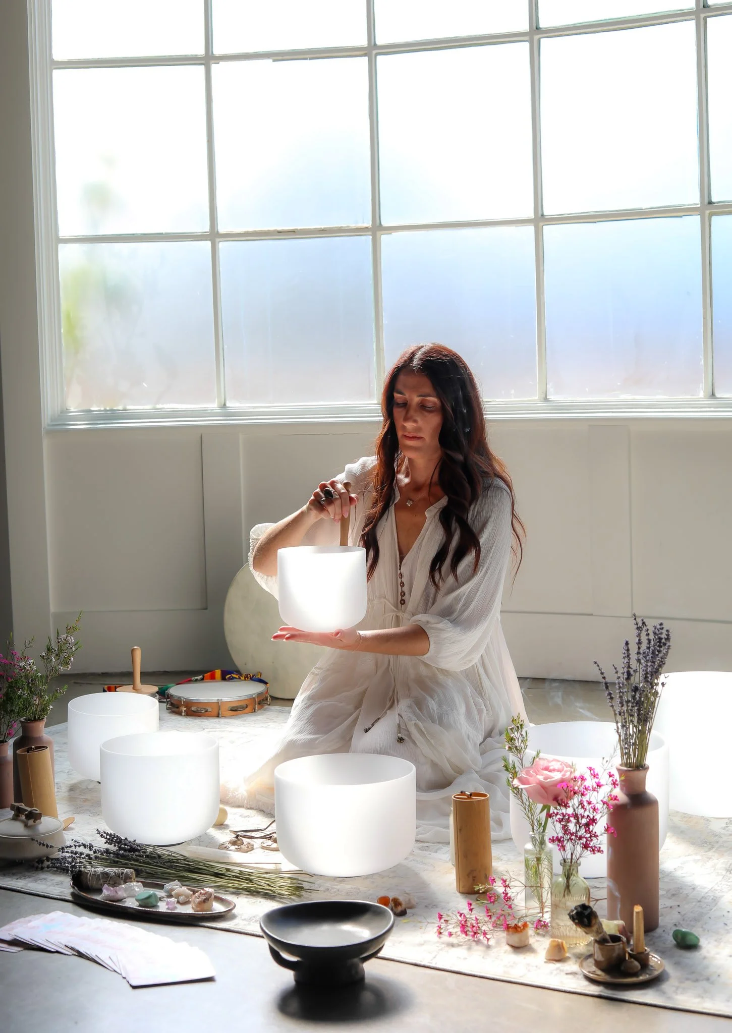A woman in a white dress kneeling on the floor surrounded by white singing bowls, plants, and flowers in a room with large windows and abundant natural light.