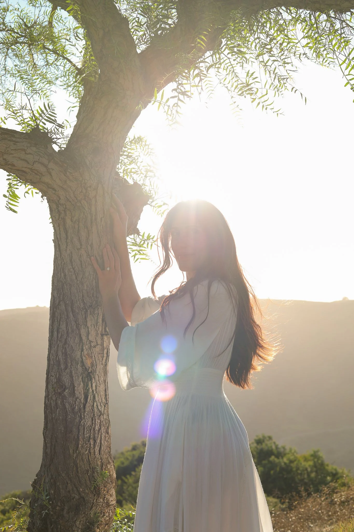 A woman with long hair, wearing a white dress, standing outdoors next to a tree with green leaves. The sun is shining brightly, creating a backlit effect and lens flare. The background shows a landscape with hills and vegetation.