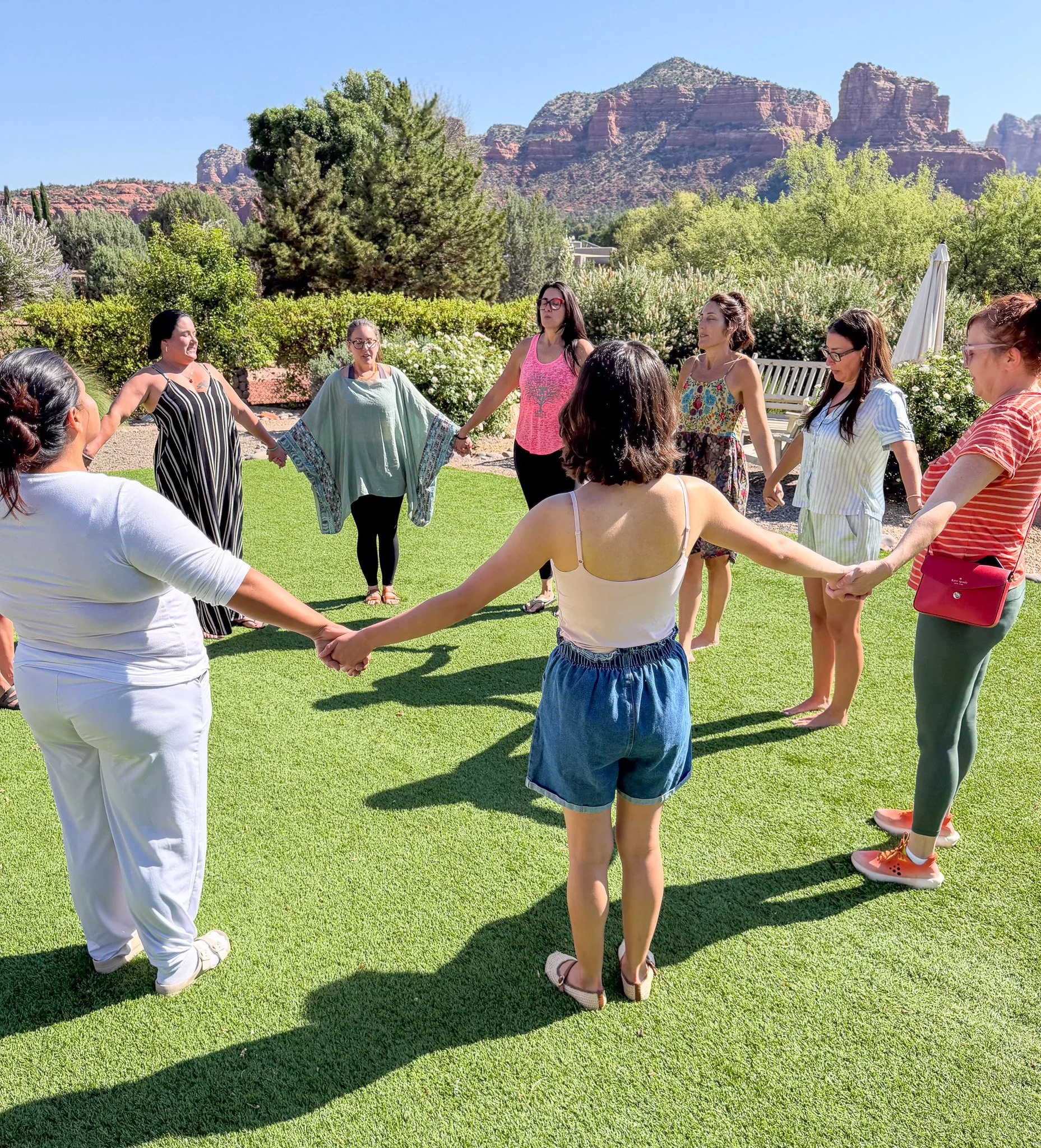 A group of women holding hands in a circle outdoors on a sunny day, with mountains and green trees in the background.