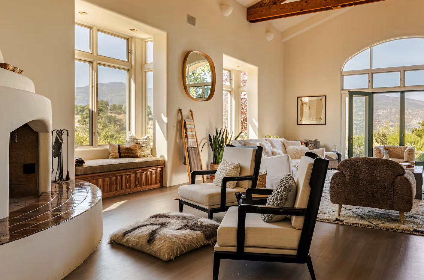 Bright living room with large windows, white walls, and a cozy seating area featuring modern and rustic furniture, plants, and a fireplace.