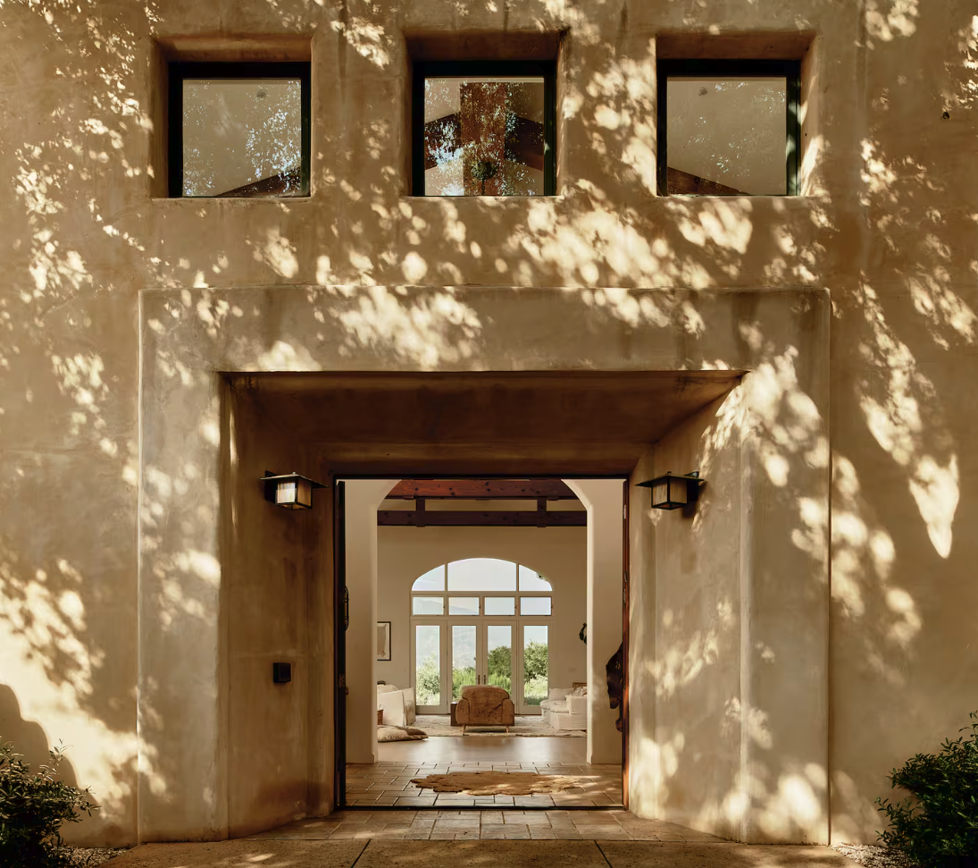 View of a Mediterranean-style house exterior with an open doorway leading to a living room, with sunlight filtering through tree leaves creating dappled shadows on the wall.