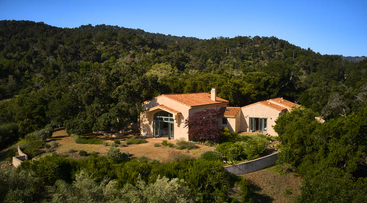 A house with a red tile roof surrounded by trees and greenery on a hillside, under a clear blue sky.
