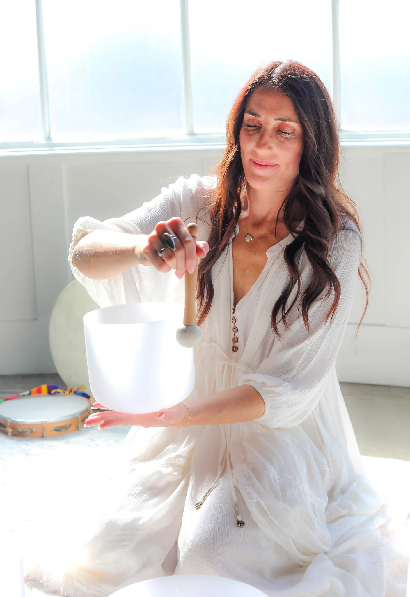 A woman with long dark hair wearing a white dress, sitting indoors near a big window, holding a glowing white container, with a tambourine and a small drum on the floor nearby.