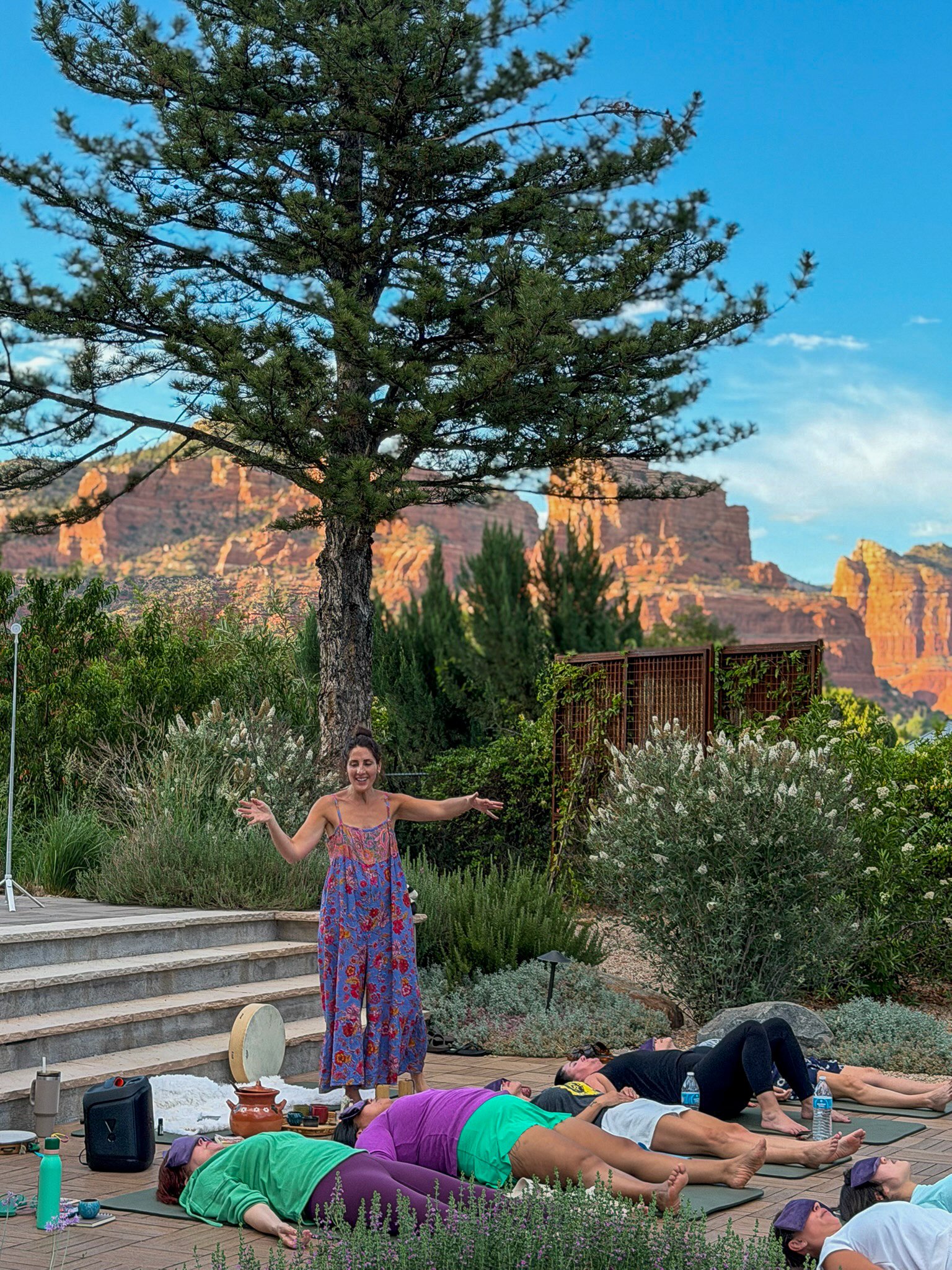 A group of people participating in a yoga class outdoors on a wooden deck, with mountains and trees in the background. Some are lying on mats, and a woman in a colorful dress is standing, leading the session with her arms outstretched.